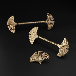 May include: Gold-colored Ginkgo leaf-shaped drawer pulls and a cabinet knob. The drawer pulls have a textured, fan-shaped design on each end, connected by a twisted bar. The cabinet knob has a similar fan-shaped design. The hardware is set against a black background.