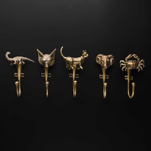 May include: A set of five brass-toned wall hooks, each featuring a different animal figure at the top. The designs include a long-necked dinosaur, a fox head, a standing cat, a ram head with curved horns, and a crab with extended claws.