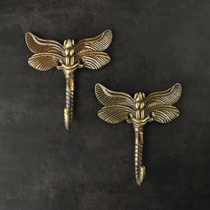 May include: Two antique brass dragonfly hooks. Each hook features detailed wings and a curved hook at the bottom. The dragonfly design adds a touch of nature-inspired decor. Perfect for hanging coats, keys, or other small items.