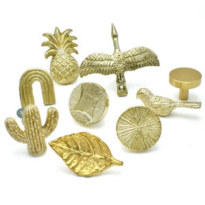 Op de afbeelding: Een set van tien gouden kastknoppen met verschillende ontwerpen, waaronder een ananas, een cactus, een blad, een vogel, een zon en een boog.