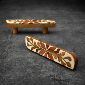 May include: Two wooden drawer pulls with intricate floral patterns inlaid in contrasting wood. The pulls are a light brown colour with a white background.