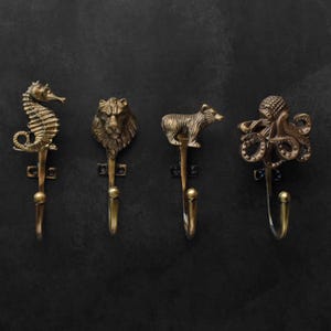 Peut inclure: Quatre crochets muraux en laiton antique avec des motifs d'animaux : un hippocampe, une t&ecirc;te de lion, un ours et une pieuvre. Chaque crochet a une figurine d'animal d&eacute;corative en haut et un crochet incurv&eacute; en dessous. Les crochets sont mont&eacute;s sur un fond sombre.