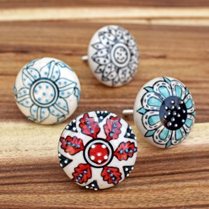 May include: Four decorative ceramic cabinet knobs with floral designs. The knobs feature white backgrounds with hand-painted patterns in teal, black, red, and blue. Each knob has a metal stem for mounting.
