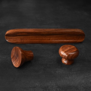 May include: A set of three wooden furniture hardware pieces. The set includes two knobs and a long, curved handle. The wood has a rich, dark brown color with visible grain patterns, set against a dark background.