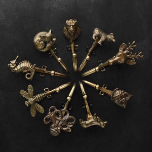 Peut inclure: Ensemble de crochets d&eacute;coratifs en laiton, chacun orn&eacute; d'un motif diff&eacute;rent : hippocampe, renard, lion, ours, cerf, hibou et libellule. Les crochets sont dispos&eacute;s en cercle sur un fond sombre.