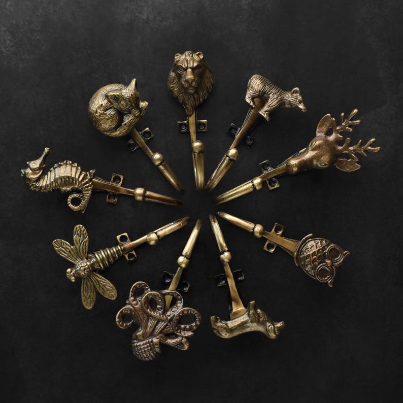 Peut inclure: Ensemble de crochets d&eacute;coratifs en laiton, chacun orn&eacute; d'un motif diff&eacute;rent : hippocampe, renard, lion, ours, cerf, hibou et libellule. Les crochets sont dispos&eacute;s en cercle sur un fond sombre.