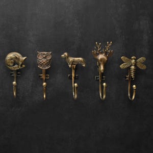 Peut inclure: Cinq crochets muraux d&eacute;coratifs en m&eacute;tal de couleur bronze, chacun avec un design animalier diff&eacute;rent. Les crochets repr&eacute;sentent un renard, un hibou, un teckel, un cerf et une abeille. Ils sont dispos&eacute;s en rang&eacute;e sur un fond sombre.