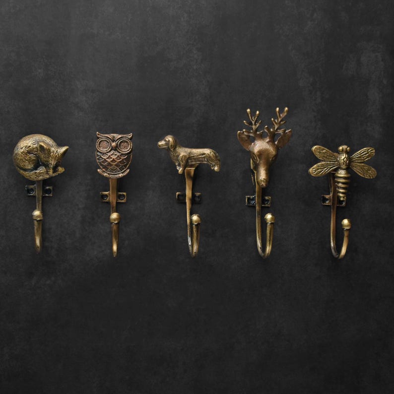 Peut inclure: Cinq crochets muraux d&eacute;coratifs en m&eacute;tal de couleur bronze, chacun avec un design animalier diff&eacute;rent. Les crochets repr&eacute;sentent un renard, un hibou, un teckel, un cerf et une abeille. Ils sont dispos&eacute;s en rang&eacute;e sur un fond sombre.