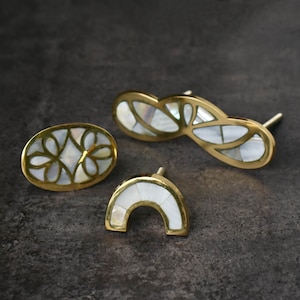 May include: Three decorative cabinet knobs with brass frames and inlaid mother-of-pearl designs. The knobs include an oval shape, a figure-eight shape, and a curved, arched shape. The knobs are set against a dark grey background.