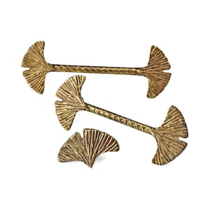 Może przedstawiać: Trzy złote metalowe przedmioty dekoracyjne. Dwa są długie, z liściastym wzorem na każdym końcu i teksturowanym środkiem. Trzeci to pojedynczy liściasty wzór. Wszystko na białym tle.