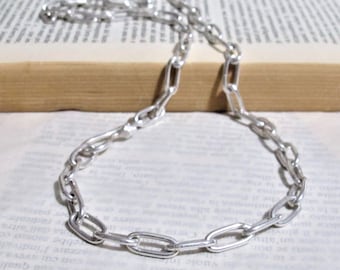 Può includere: Una collana a catena d'argento con uno stile di maglia che ricorda una graffetta. La catena è appoggiata su un libro.