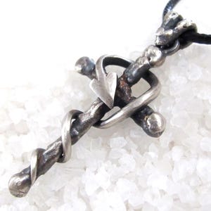 May include: A silver-toned cross pendant with a heart accent, suspended on a black cord. The cross has a textured, rustic appearance, with a heart shape at the intersection of the crossbars. The design is detailed and intricate, suggesting handcrafted artistry.