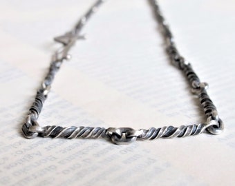 Può includere: Una collana con catena d'argento dal design intrecciato. La catena è composta da piccoli pezzi di metallo collegati tra loro e intrecciati per creare un look unico.