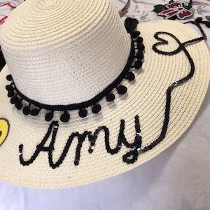 May include: A cream-colored sun hat with a black pom-pom trim and the name "Amy" spelled out in black sequins. A yellow smiley face is on the brim. The hat is decorated with black sequins and a heart design.