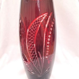 Puede incluir: Un jarrón de cristal rojo rubí con la parte superior acampanada y un diseño floral detallado. El jarrón presenta patrones grabados y un borde festoneado. La base del jarrón es de un tono rojo más oscuro.