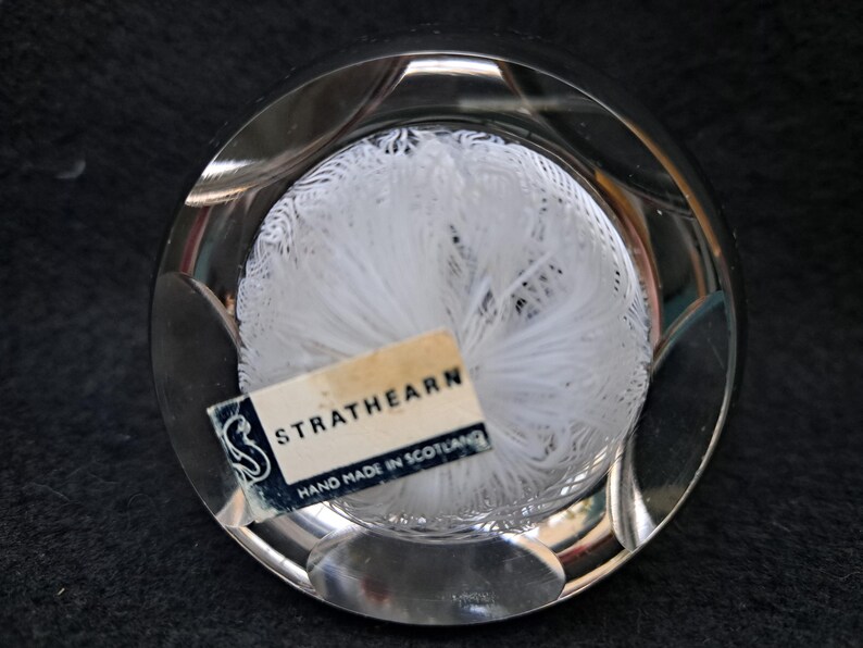 May include: A clear glass paperweight with a white, feathery design inside. The paperweight has a label that reads "Strathearn Hand Made in Scotland."