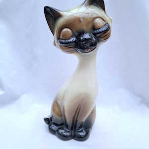 May include: A vintage ceramic Siamese cat figurine. The cat has closed eyes, black ears, and a black nose. The cat's fur is a mix of tan, brown, and cream colors. The cat is sitting upright with its paws tucked beneath it.