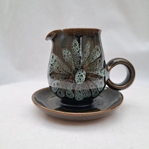 May include: A ceramic creamer and saucer set. The creamer is dark brown with a floral pattern in shades of green and white. The saucer is a matching dark brown. The creamer has a handle and a spout.