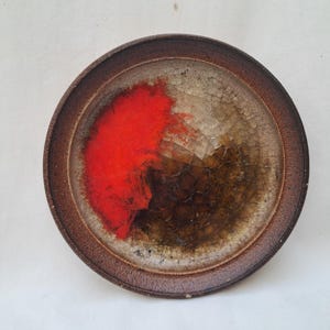 Peut inclure: Une assiette en céramique circulaire avec un bord brun foncé et un centre à finition émaillée craquelée. La surface présente une éclaboussure abstraite rouge vif contrastant avec un dégradé de tons beige, ocre et brun profond.