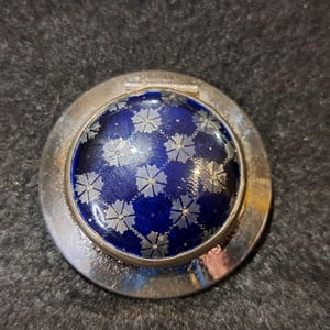 Könnte beinhalten: Ein silberfarbener Kompaktspiegel mit einer dunkelblauen, floral gemusterten Oberfläche. Das Design zeigt kleine, silberfarbene Blütenformen auf einem dunkelblauen Hintergrund. Der Kompaktspiegel hat einen Klappdeckel und eine runde Form.