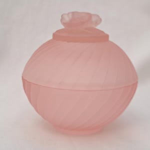 May include: A frosted pink glass jar with a spiral design and a flower-shaped lid. The jar is round and has a matte finish, giving it a soft, delicate appearance. The lid sits atop the jar, completing the elegant design.