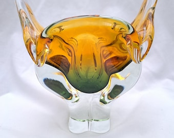 May include: A clear glass bowl with a unique, abstract design. The bowl is amber and green in colour and has two large, curved handles.