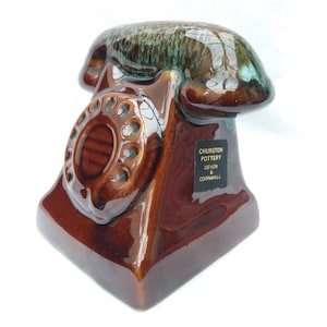 Może przedstawiać: Ceramiczna figurka telefonu w kolorze brązowym z zielonym szkliwem. Figurka ma tarczę obrotową i etykietę z napisem "Churston Pottery Devon & Cornwall".