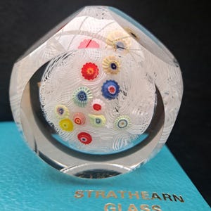 May include: A clear glass paperweight with a white swirl design and colorful flowers inside. The paperweight is on a blue box with the text "Strathearn Glass".