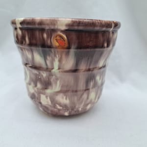 May include: A ceramic plant pot with a distinctive, textured pattern. The exterior displays a mix of brown, cream, and light green shades. The pot has a slightly flared rim and a rounded base. A small sticker is visible on the side.