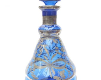 May include: A cobalt blue glass decanter with a silver-colored rim and stopper. The decanter features a floral design in gold. The stopper is cylindrical and the body of the decanter is pear-shaped.