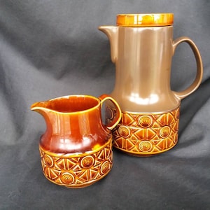 Könnte beinhalten: Ein braunes und oranges Keramik-Kaffee- und Milchkännchen-Set mit geometrischem Muster. Die Kaffeekanne hat einen Deckel und einen Griff. Das Milchkännchen hat einen Griff.