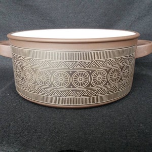 Könnte beinhalten: Eine braune Keramik-Auflaufform mit weißem Innenraum und einem geometrischen Muster in Gold. Das Muster zeigt Kreise mit Speichen und Dreiecke.