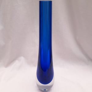 Puede incluir: Un jarrón alto y elegante con una base clara y redondeada y una sección superior azul vibrante. El jarrón tiene una forma esbelta y alargada, que transiciona de transparente a azul profundo. El vidrio parece liso y reflectante, mostrando un diseño moderno.