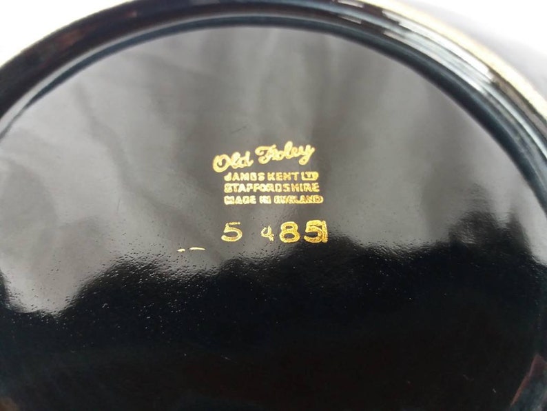May include: Close-up of the bottom of a black ceramic plate with gold lettering. The text reads "Old Foley, James Kent Ltd, Staffordshire, Made in England, 5 4851".