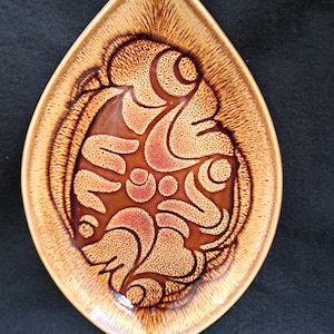 May include: A vintage, leaf-shaped ceramic dish with a brown and tan glaze. The dish features a central design of stylized floral or abstract shapes in shades of brown and red. The dish is approximately 25 cm long.