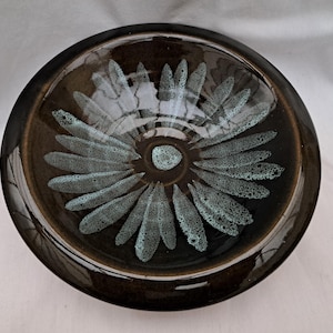 May include: A round, ceramic bowl with a dark brown exterior and a floral design in the center. The flower design features teal-colored petals radiating from a central point. The bowl has a glossy finish.
