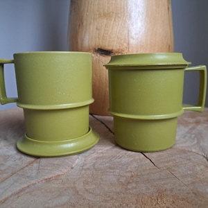 Puede incluir: Dos tazas de plástico verde oliva con asas y una tapa a juego. Las tazas tienen un diseño único con una apariencia apilada. Las tazas están sobre una superficie de madera, con un objeto de madera en el fondo.