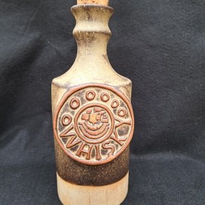 Puede incluir: Botella de whisky de cerámica con tapón de corcho. La botella es de color marrón y beige con un emblema circular que dice "Whisky". El emblema presenta un diseño de barco. La botella tiene una base de color beige claro.