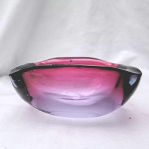Puede incluir: Un pequeño cuenco de cristal redondo con un degradado de colores vivos que va desde un magenta intenso y saturado en el borde superior hasta un tono púrpura más claro y translúcido en la base. El cuenco tiene un borde liso, grueso y pulido.