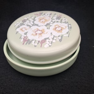 May include: A two-piece, pale green ceramic container. The lid features a floral design in white, tan, and grey. The container is round and appears to be a trinket box or small storage dish.