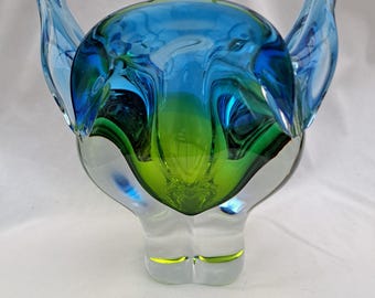 May include: A decorative glass ornament with a distinctive form. It showcases blue and green hues, complemented by clear glass details. The design incorporates upward-curving features, resulting in an intriguing shape. The glass has a polished surface.