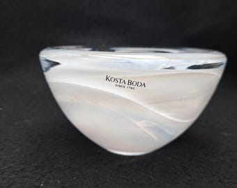 May include: A Kosta Boda glass bowl featuring a smooth, rounded shape with swirling white and translucent patterns throughout the body. The brand name Kosta Boda Since 1742 is printed in black text on the upper exterior surface of the glass.