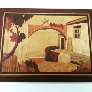 Può includere: Un'opera d'arte murale in legno che rappresenta una scena di una casa con un arco d'ingresso, un albero e una montagna in lontananza. L'opera d'arte è realizzata con diversi tipi di legno, creando un effetto a mosaico.