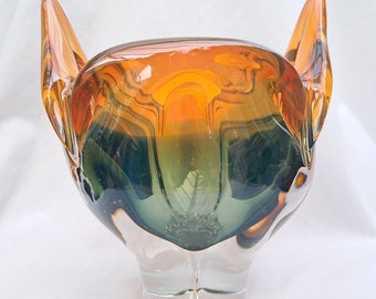 May include: A glass bowl with a unique, organic shape. The bowl is made of clear glass with a layered effect of orange, green, and brown.
