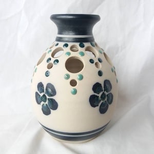 Puede incluir: Un jarrón de cerámica blanco con cuerpo redondo y borde negro. El jarrón tiene un patrón decorativo de flores azules y puntos verdes. Hay agujeros en el jarrón para los tallos de las flores.