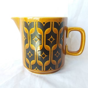 以下が含まれることがあります： 取っ手と注ぎ口が付いたセラミックピッチャー。ピッチャーは温かみのある茶色で、黒と金の幾何学模様が繰り返されています。ハンドルは同じ茶色です。