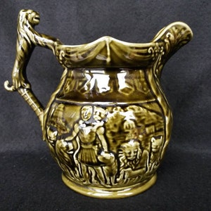 Puede incluir: Jarra de cerámica con un esmalte verde oliva oscuro. El asa está esculpida con una cabeza y una pata de león. El cuerpo presenta escenas en relieve de figuras y animales, probablemente de una narrativa clásica. La jarra tiene un pico decorativo.