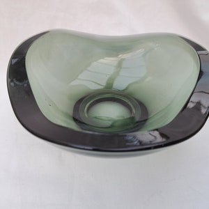 Puede incluir: Un cuenco de vidrio verde salvia en forma de corazón con un borde gris oscuro. El cuenco tiene un acabado liso y brillante y una hendidura circular en el centro. El diseño es simple y elegante.