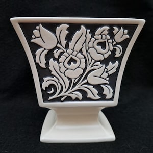 Könnte beinhalten: Eine weiße Keramikvase mit quadratischem Sockel und ausgestellter Oberseite. Die Vase hat eine schwarze Tafel mit einem weißen floralen Reliefdesign. Das florale Design umfasst Rosen, Blätter und andere botanische Elemente. Die Vase dient wahrscheinlich dekorativen Zwecken.