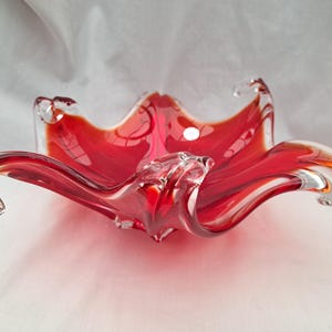 May include: A red glass bowl with a wavy, organic design. The bowl is made of clear glass with a red tint and has a unique, flowing shape.
